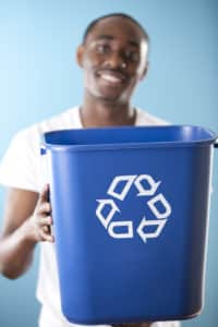 An African American young adult man holds out a recycling bin. Selective focus is on the recycling bin.